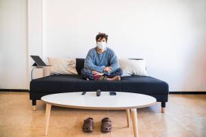 An image showing a person sitting on a couch wearing a mask, with slippers on the floor and a coffee table in front of them